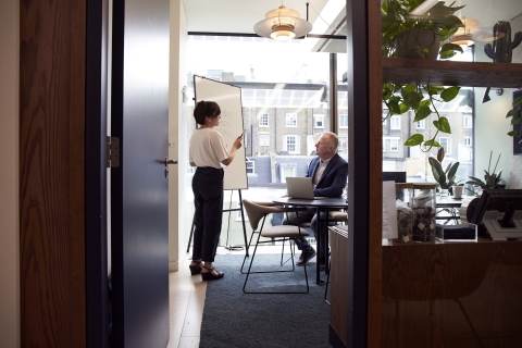 Person standing next to a whiteboard talking to a man who is seated